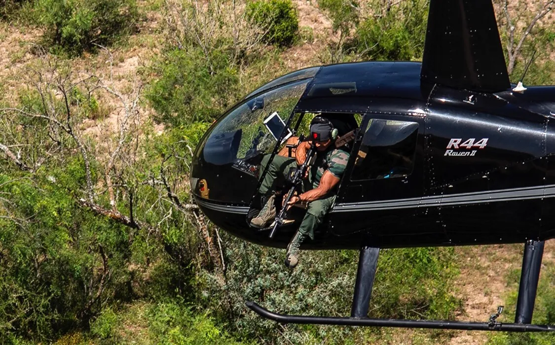 A shooter leaning out of a helicopter in low flight over the Texas brush