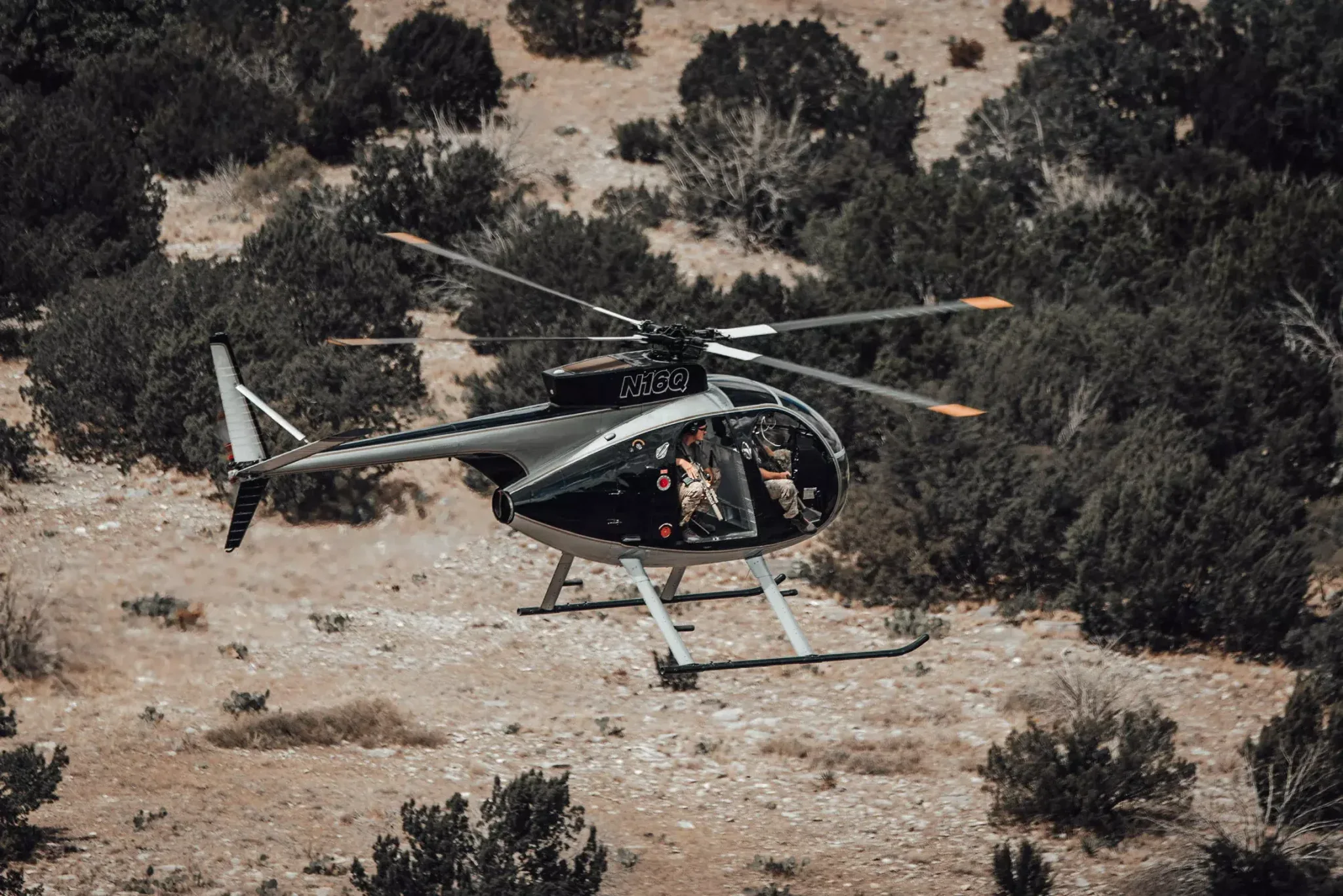 A hunting helicopter in low flight over West Texas scrub, a shooter seated in the open doorway